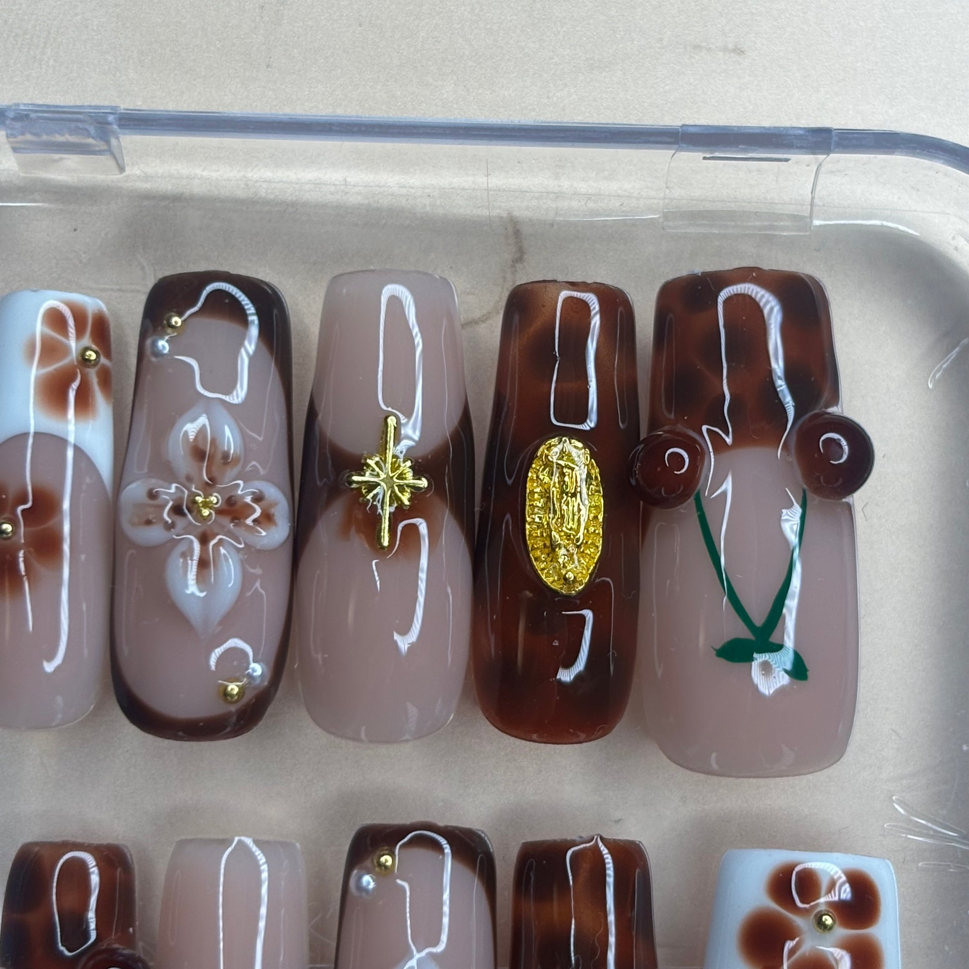 A set of decorative press-on nails displayed in a clear plastic tray. The nails feature various designs including brown and beige marbled patterns, white line art, floral motifs, gold star and oval charms, and green leaf details.